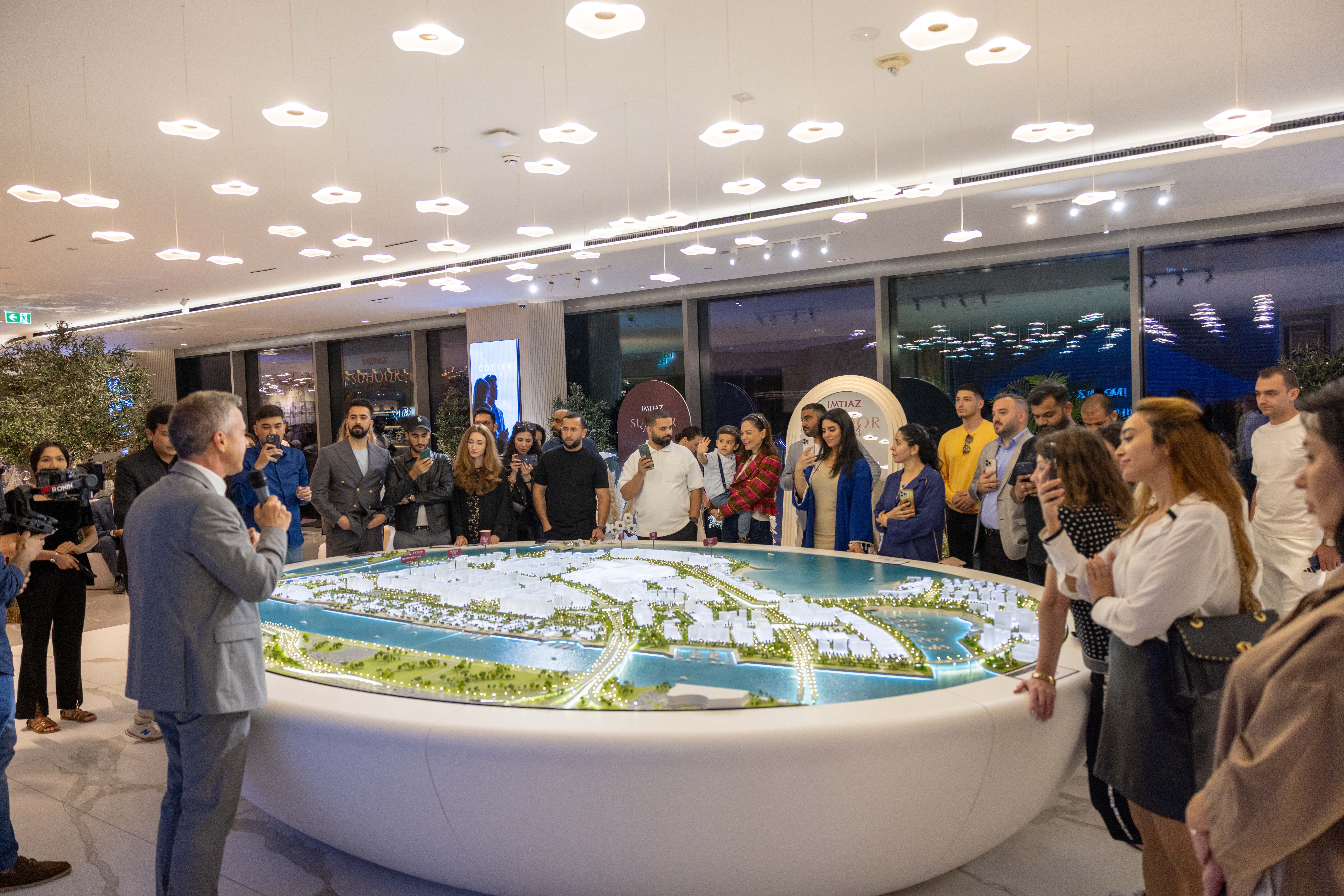 Events?Page=3 A group of people attentively watches a presentation beside a detailed architectural model of a coastal development.
